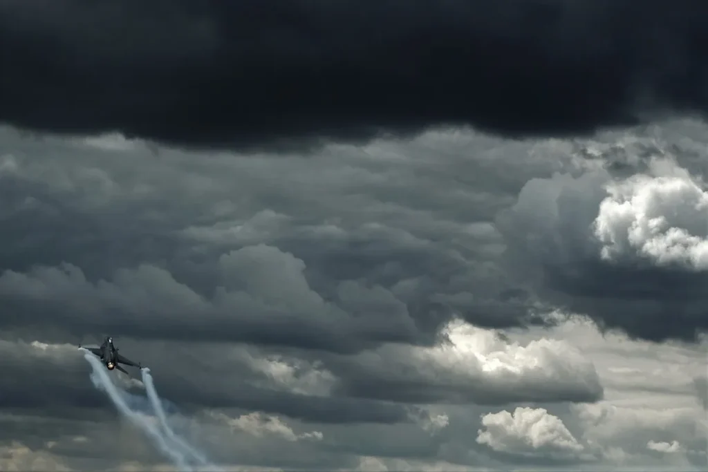 Avião caça militar soltando fumaça enquanto voa sob nuvens escuras de tempestade no céu.