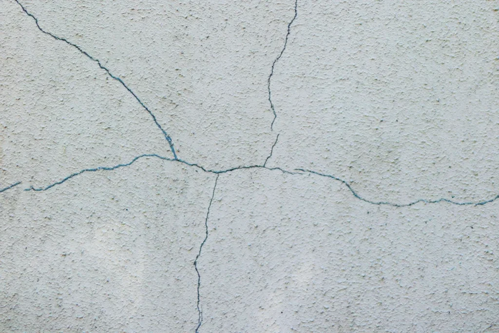 Fissuras finas e ramificadas em parede com reboco texturizado de cor clara.