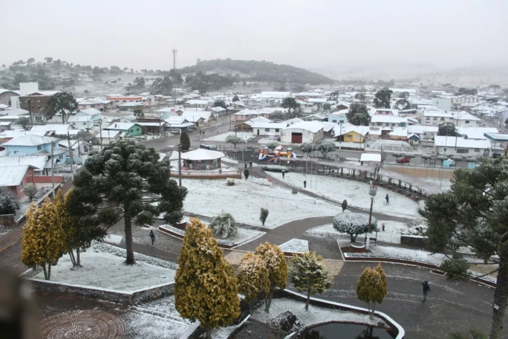 Vista panorâmica de uma cidade brasileira com casas, ruas e árvores cobertas pela neve durante o inverno.