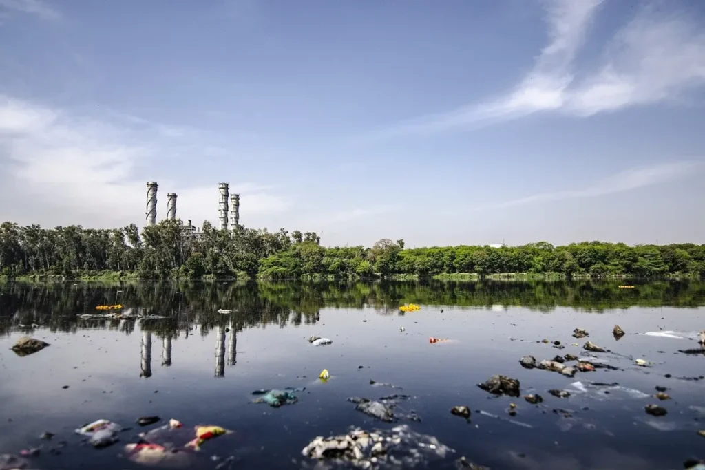 Rio poluído com lixo e resíduos industriais flutuando na superfície, próximo a área de fábrica com chaminés e árvores ao fundo.