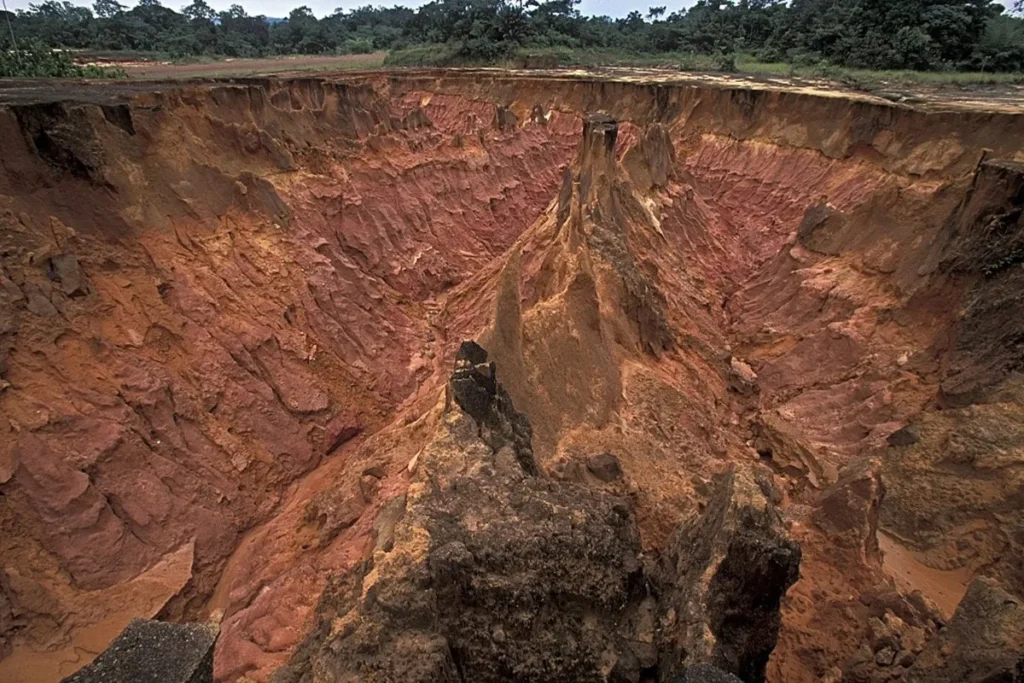 Grande voçoroca formada por erosão hídrica em solo vermelho, com paredes íngremes e profundas, cercada por vegetação ao fundo.