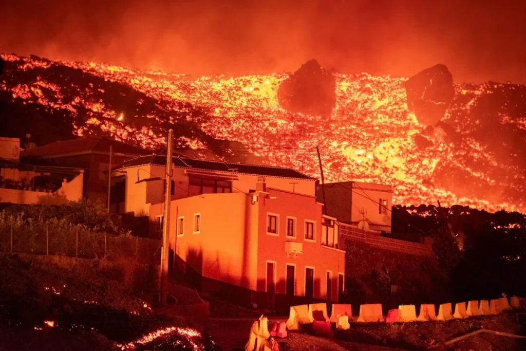 Fluxo de lava incandescente descendo de um vulcão e avançando em direção a casas iluminadas pelo brilho do magma
