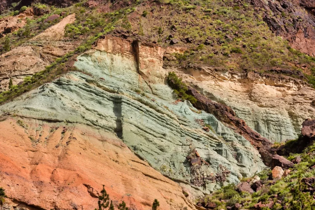 Parede rochosa com camadas de cores diferentes entre vermelho, verde e bege, coberta por vegetação em algumas áreas