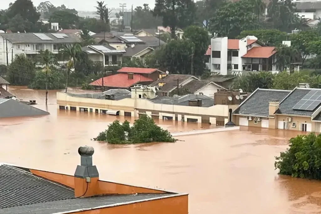 Enchente em bairro residencial com ruas e casas parcialmente submersas pela água barrenta após fortes chuvas, evidenciando os impactos das inundações urbanas.