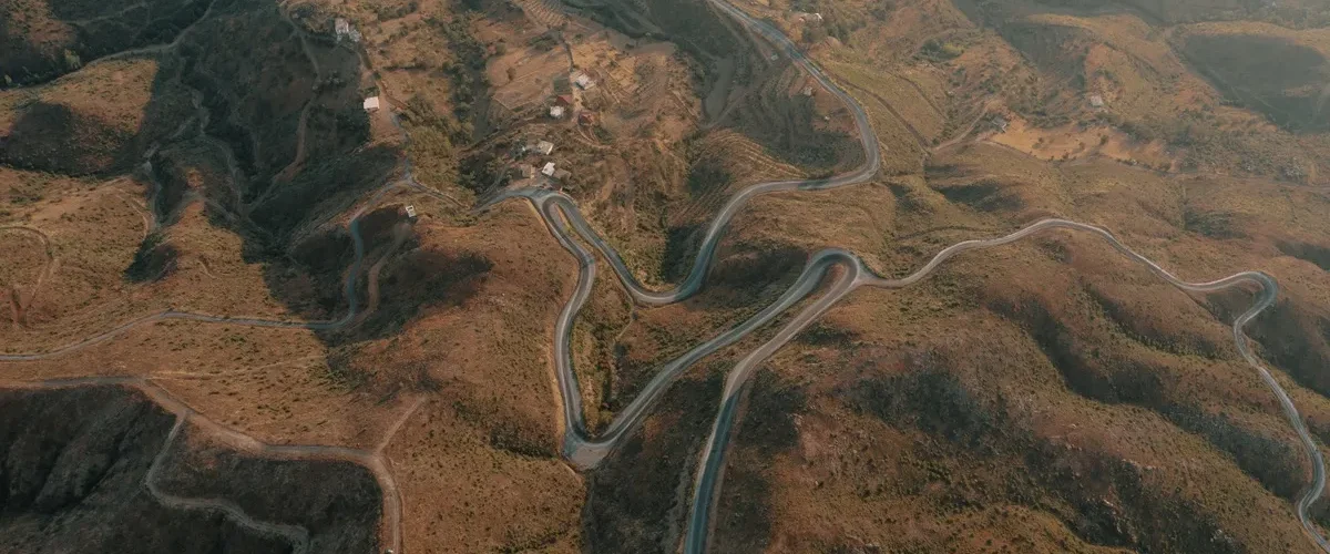 Vista aérea de estrada sinuosa em meio a montanhas áridas, cercada por pequenas casas e áreas de vegetação.