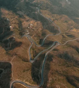 Vista aérea de estrada sinuosa em meio a montanhas áridas, cercada por pequenas casas e áreas de vegetação.