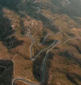 Vista aérea de estrada sinuosa em meio a montanhas áridas, cercada por pequenas casas e áreas de vegetação.
