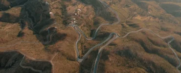 Vista aérea de estrada sinuosa em meio a montanhas áridas, cercada por pequenas casas e áreas de vegetação.
