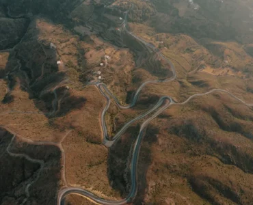Vista aérea de estrada sinuosa em meio a montanhas áridas, cercada por pequenas casas e áreas de vegetação.