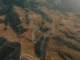 Vista aérea de estrada sinuosa em meio a montanhas áridas, cercada por pequenas casas e áreas de vegetação.
