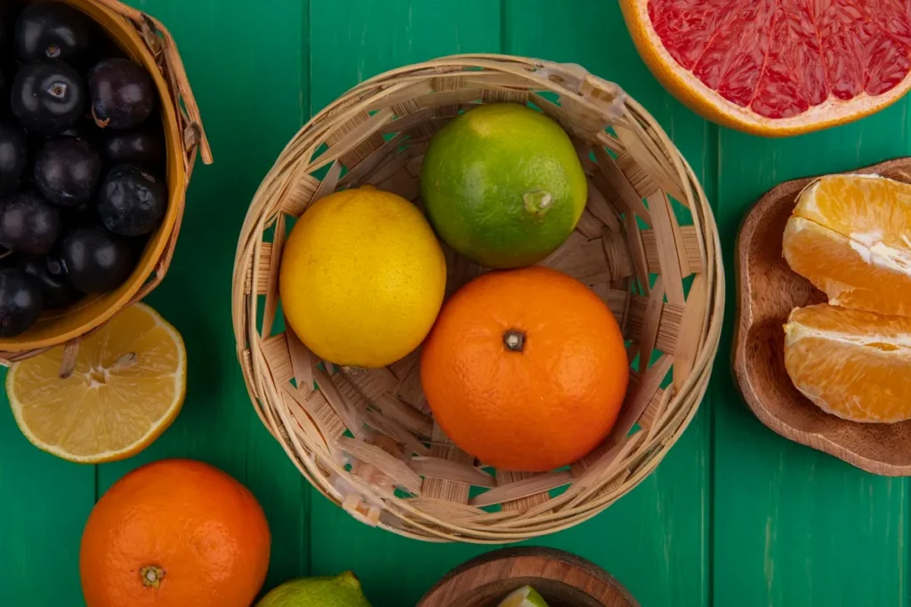 Cesta de palha com frutas cítricas como laranja, limão e lima sobre mesa verde, acompanhadas de uvas pretas e fatias de grapefruit e laranja ao redor.