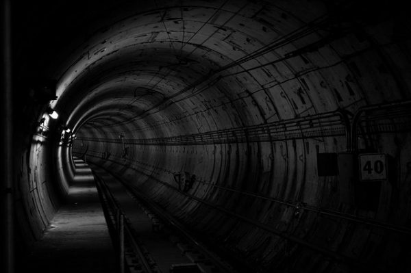 Imagem de um túnel iluminado por uma lâmpada à esquerda, com trilhos no chão.