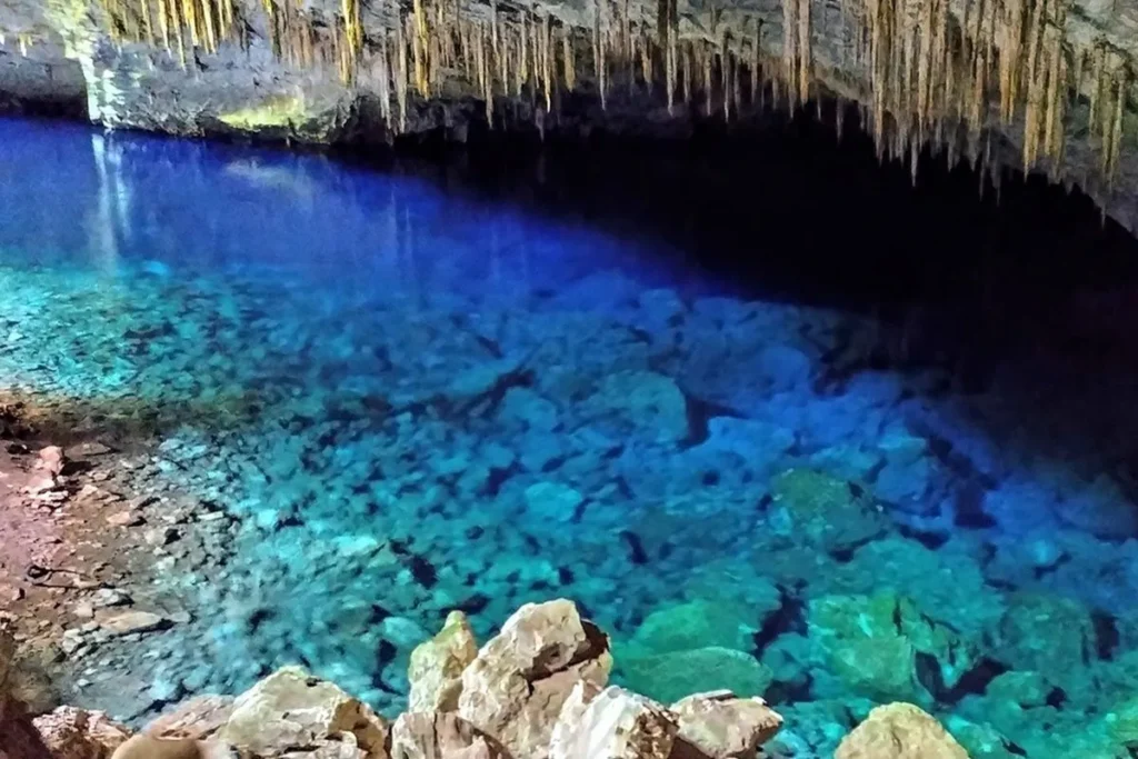 Interior de caverna com águas azul-turquesa cristalinas, estalactites no teto e rochas submersas visíveis.