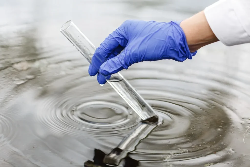 Mão com luva azul segurando tubo de ensaio para coleta de amostra de água em lago ou rio para análise de salinidade e qualidade.