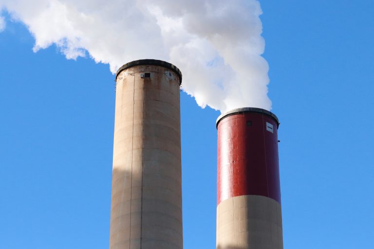 Foto de fumaça saindo de duas torres em uma fábrica, despontando para o ar, para ilustrar os impactos da poluição atmosférica.