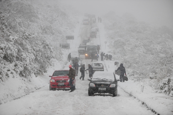 Foto de uma estrada cheia de neve, com carros parados cobertos de neve, e pessoas levando malas.