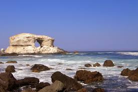 Imagem de uma estrutura rochosa ao horizonte em uma praia, com ondas e pedras espalhadas.