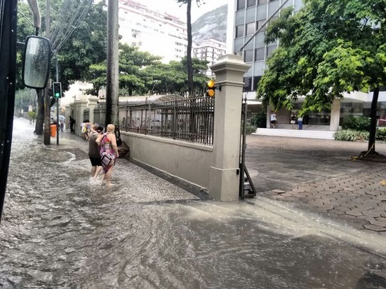 Foto de um ponto de alagamentos no Rio de Janeiro, com a calçada parcialmente encoberta por água e duas mulheres andando com água até a canela. Não constitui enchentes ainda.