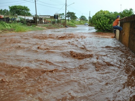 Foto de uma enxurrada no Mato Grosso do Sul, com água barrenta em movimento numa rua, e uma pessoa pendurada num muro à direita para evitar pisar na água.