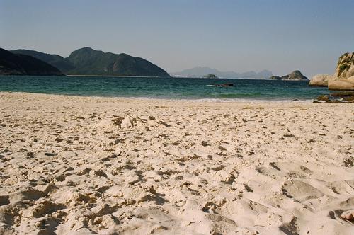 Foto de uma praia, com areia à frente, o mar atrás e, no horizonte, montanhas.