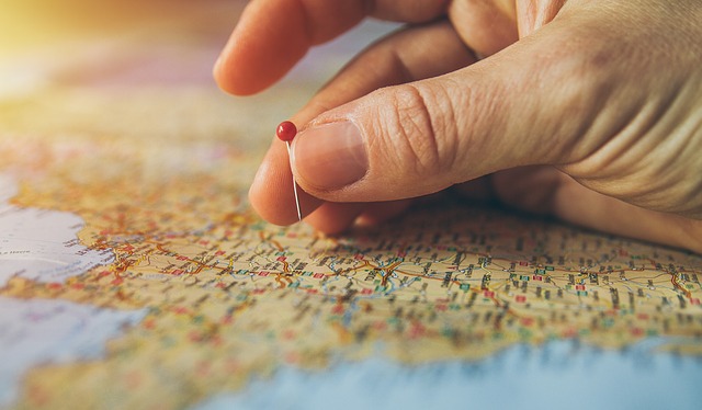 Imagem de uma mão branca segurando um pin vermelho sobre um mapa de representações cartográficas.