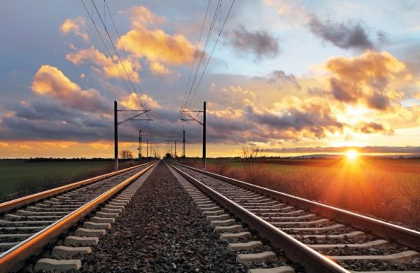 Foto de uma ferrovia com dois trilhos indo em direção ao horizonte, com o por do sol à direita e nuvens no céu.