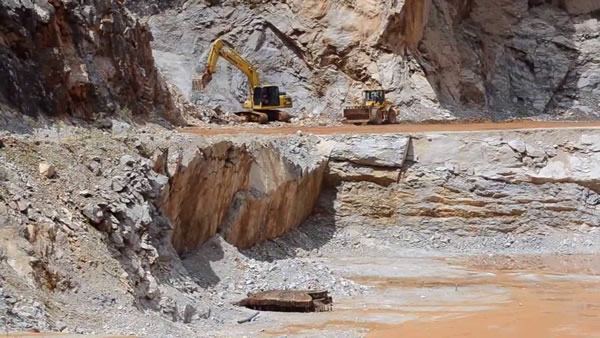 Foto de uma mina com uma escavadeira, em que se realiza o desmonte de rocha.