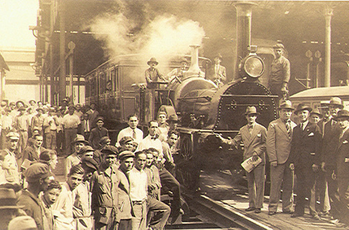Foto em sépia (amarelada) de uma locomotiva ferroviária e várias pessoas ao redor, olhando para a câmera.