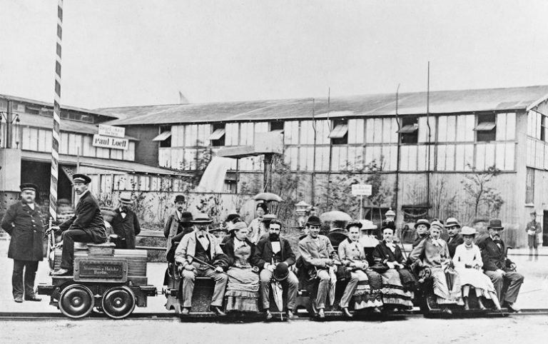 Foto preta e branca de homens e mulheres sentados no vagão de um motor de tração, um dos primeiros modelos do sistema de eletrificação ferroviária.