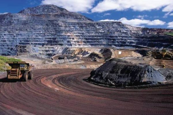 Foto de uma jazida de minérios de ferro, com um trator passando em uma estradinha de terra à esquerda.