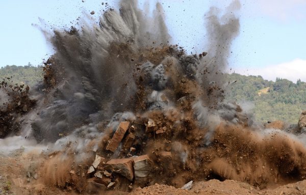 Foto da implosão de uma pedreira, com fumaça, areia e detritos voando no ar.