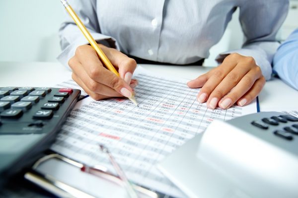 Foto de uma mulher sentada a uma mesa, dos ombros para baixo, com foco em suas mãos rascunhando com lápis em uma folha de números, representando a orçamentação.