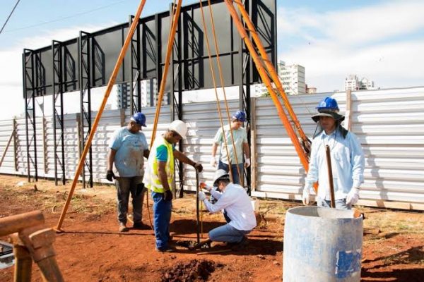 Foto de cinco operários usando equipamentos de proteção na frente de um canteiro de obras, começando o processo de sondagens do solo, após a definição dos elementos básicos da construção civil.