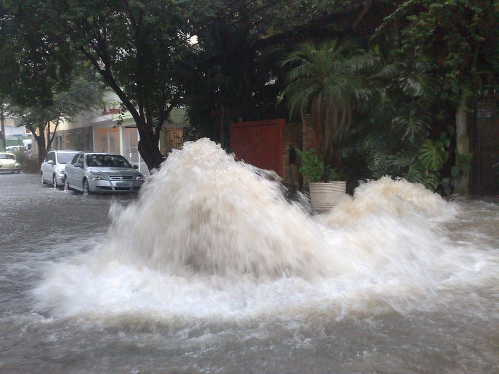Via de São Paulo inundada devido à deficiência do sistema de drenagem urbana. Na foto, uma rua completamente alagada, com jorros de água vindos do esgoto para cima.