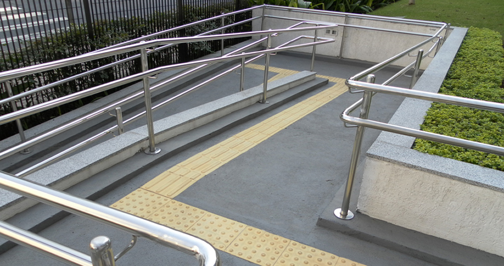 Foto de uma rampa de acesso com corrimão duplo em diferentes alturas do chão e com uma linha de piso táctil.