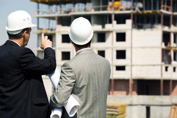 Imagem de dois homens de pé e com capacete, de costas para a foto, um do lado do outro, olhando para um prédio em construção à sua frente.