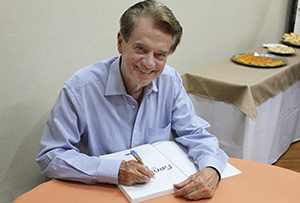 Foto do professor Nivaldo Chiossi, de blusa social, assinando um livro e sorrindo para a câmera.