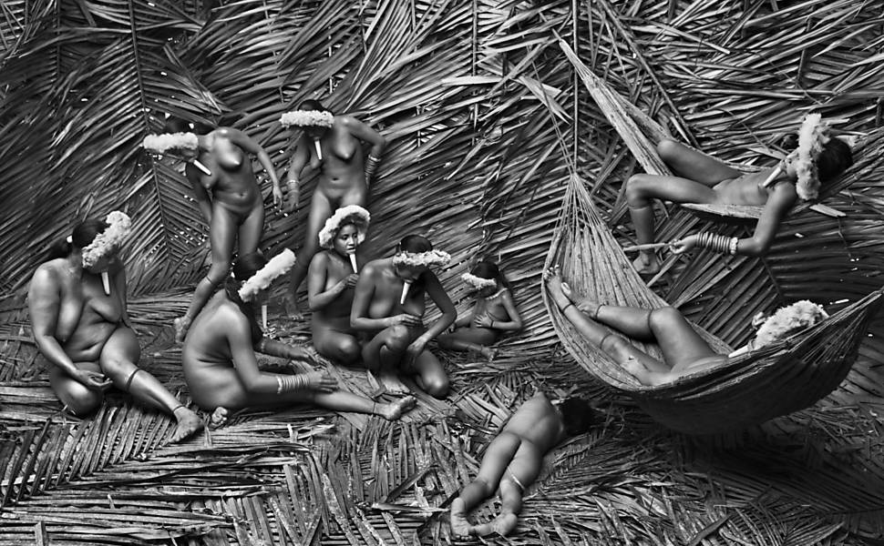 Fotografia preta e branca de indígenas nuas e com adereços na cabeça sentadas, de pé ou deitadas em redes, num chão coberto de folhas de palmeiras.