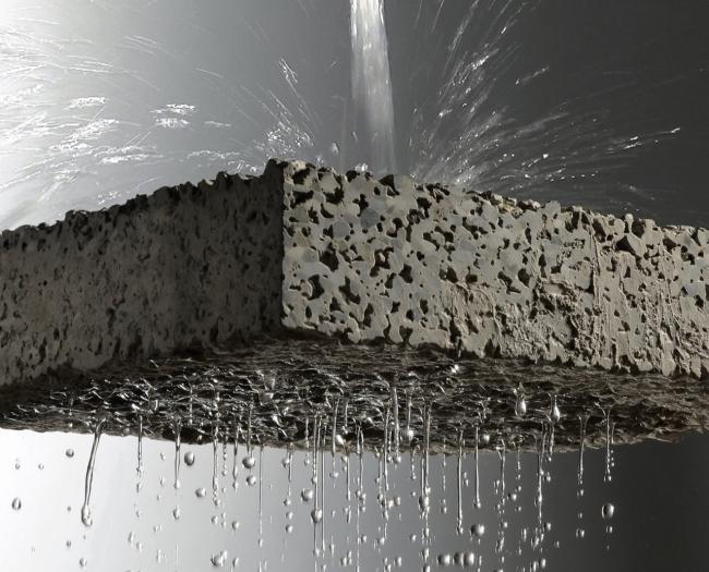 Pavimentação permeável: imagem de um pavimento poroso cinza, com água sendo jorrada em cima dele e passando por ele, pingando em gotas.