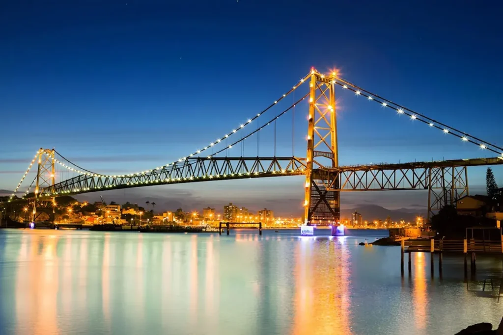 Ponte Hercílio Luz iluminada à noite, refletindo suas luzes na água, com vista para a cidade de Florianópolis.
