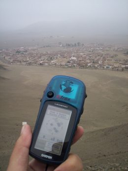 Foto de uma mão segurando um GPS, e atrás um campo de terra.