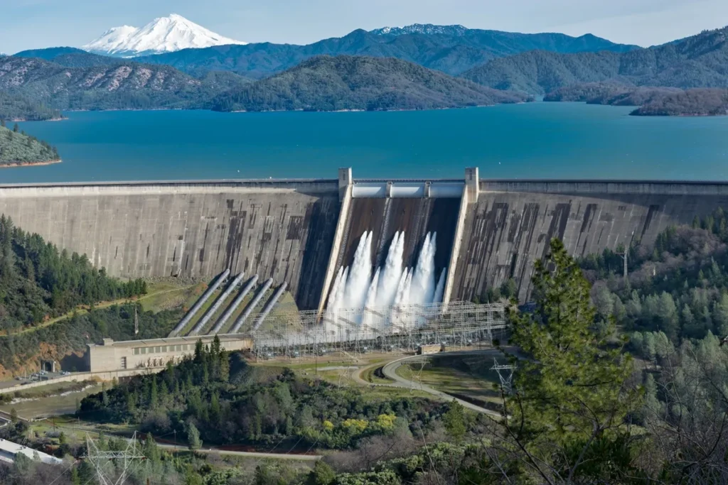 Vista panorâmica de uma usina hidrelétrica com barragem liberando água, lago ao fundo e montanhas com neve.