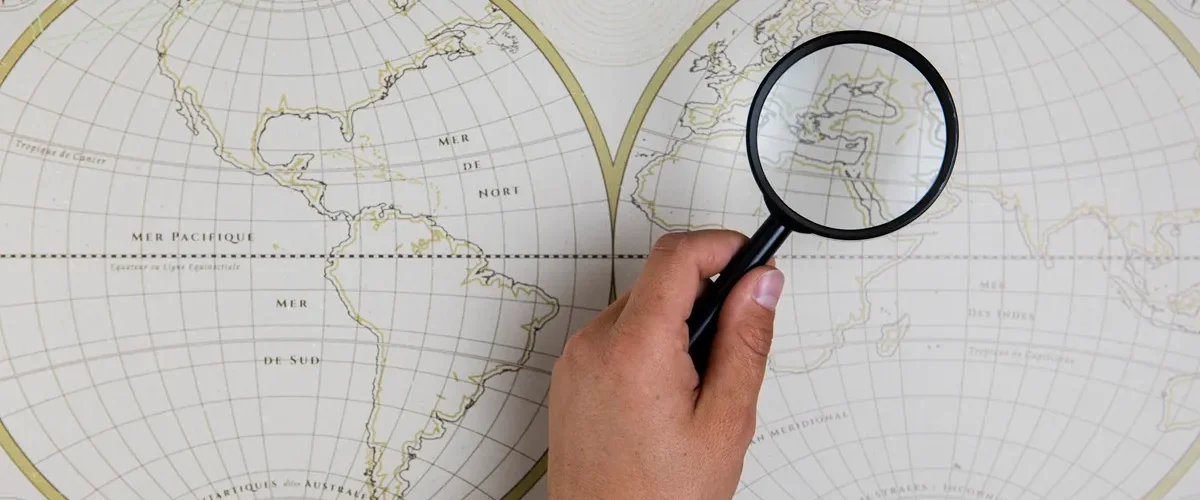 Mão segurando lupa sobre mapa mundi decorativo, ampliando a região do Mediterrâneo.