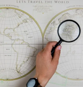 Mão segurando lupa sobre mapa mundi decorativo, ampliando a região do Mediterrâneo.