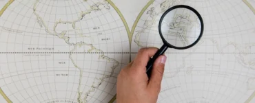 Mão segurando lupa sobre mapa mundi decorativo, ampliando a região do Mediterrâneo.