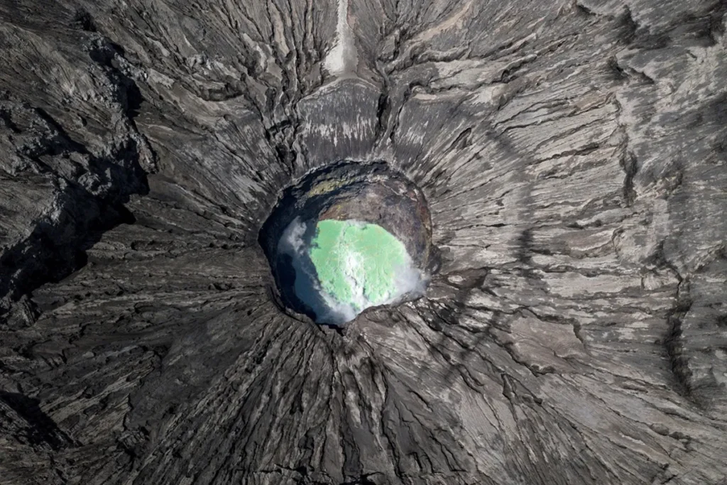Vista aérea de uma cratera vulcânica com lago esverdeado em seu interior, rodeada por rochas escuras e fissuras radiais.
