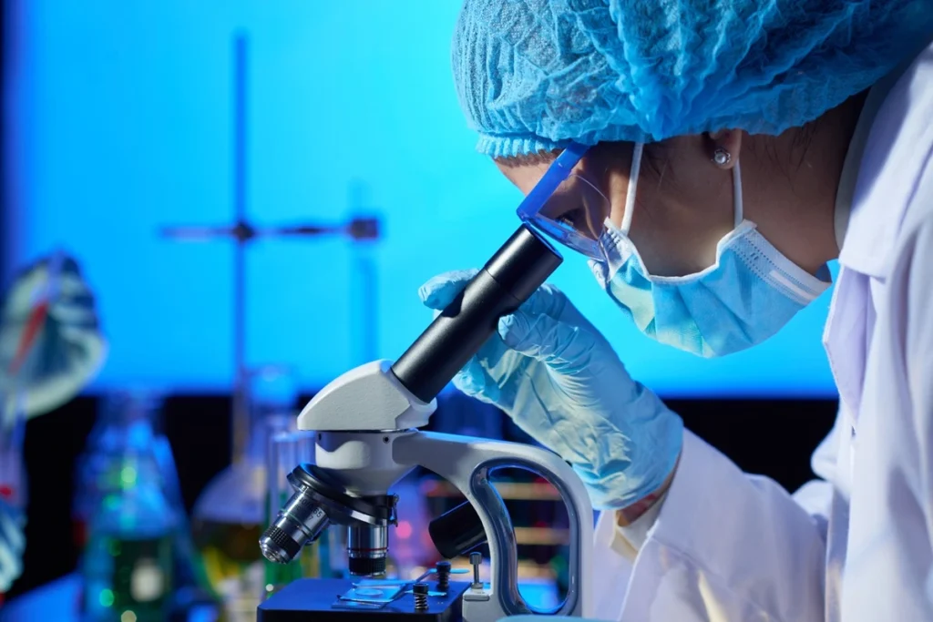 Cientista com máscara e luvas observando amostra em microscópio no laboratório