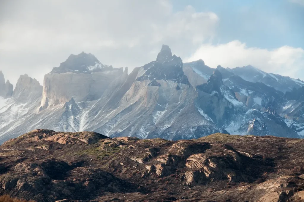 Vista panorâmica da Cordilheira dos Andes com montanhas escarpadas, picos nevados e formações rochosas sob um céu parcialmente nublado.