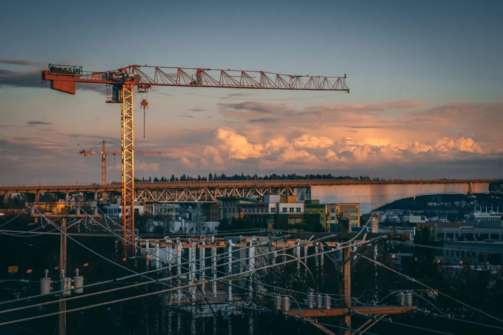 Guindaste de torre em área urbana com ponte ao fundo e céu alaranjado ao entardecer.