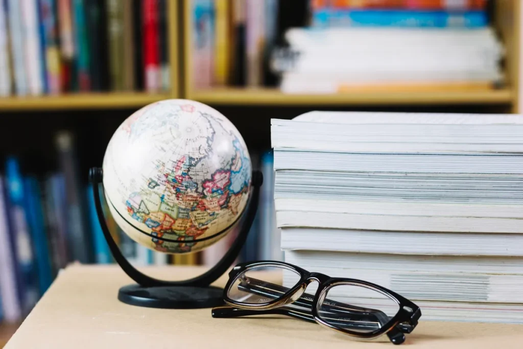 Globo terrestre sobre mesa ao lado de pilha de livros e óculos de leitura.